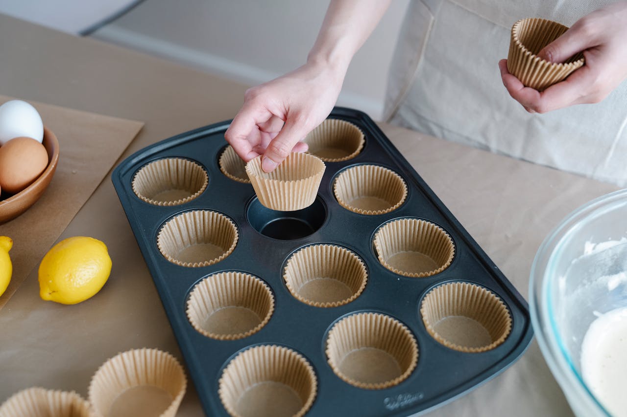 Female preparing cupcake tray with baking ingredients like eggs and lemons on kitchen counter.